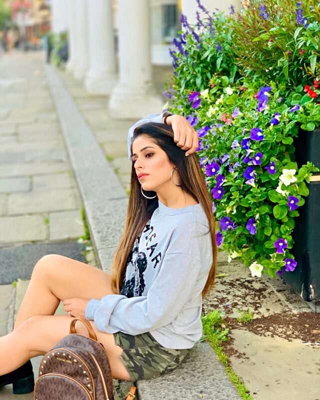 Roopsi Bansal in a grey graphic sweatshirt and camo shorts, sitting on a curb next to a wall of purple and white flowers in London