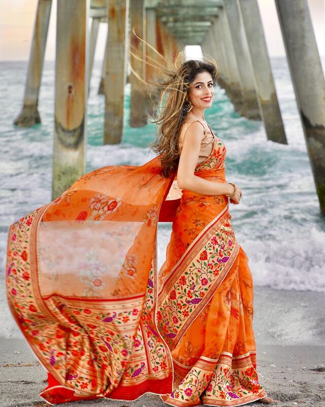 Roopsi Bansal in a flowing orange floral-bordered saree under Juno Beach Pier, hair and pallu caught in the wind, waves crashing behind her