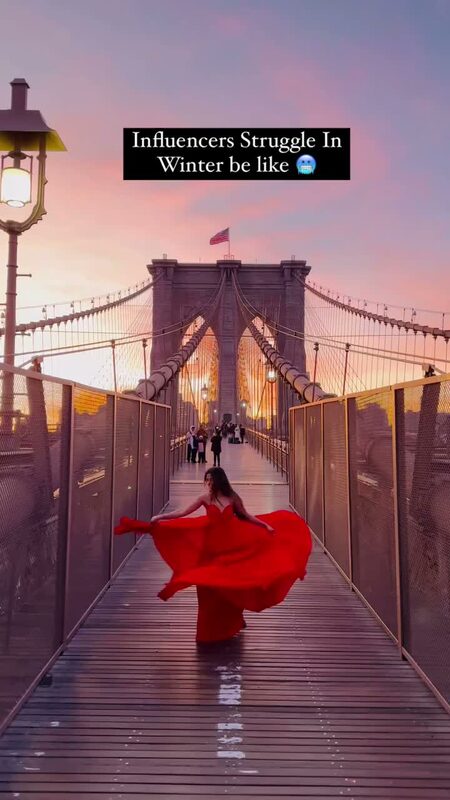 Roopsi Bansal in a flowing red gown on the Brooklyn Bridge at sunset, captioned 'Influencers Struggle In Winter be like'