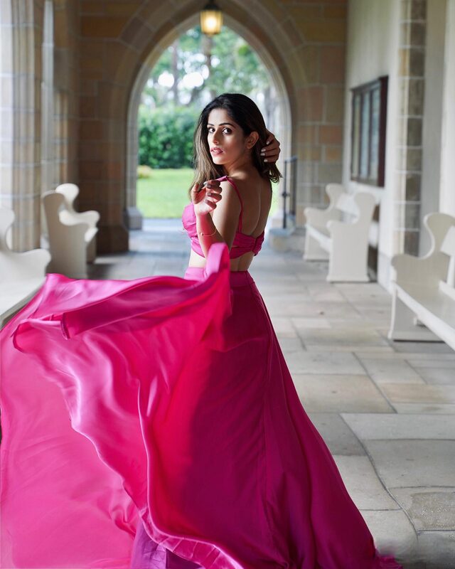 Roopsi Bansal in a hot pink two-piece satin gown, skirt swept mid-twirl, glancing back over her shoulder in a Gothic stone colonnade at Bethesda-by-the-Sea