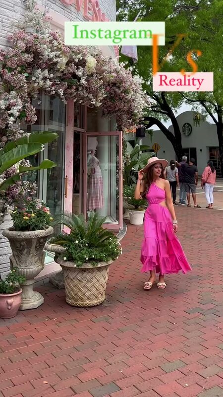 Roopsi Bansal in a hot pink tiered halter maxi dress from Shein and a wide-brim hat, walking a brick sidewalk past a flower-covered boutique storefront