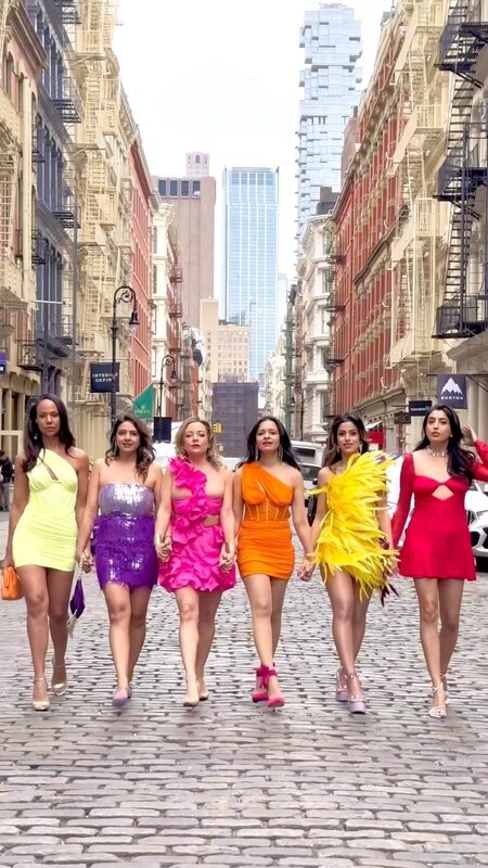 Roopsi Bansal and five other women in six different bright colored dresses walking hand-in-hand down a cobblestone SoHo street for International Women's Day