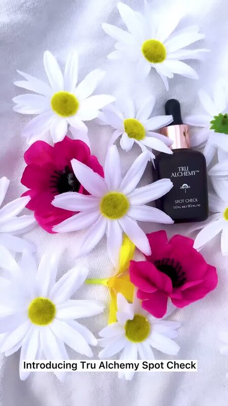 A bottle of Tru Alchemy Spot Check serum styled among white daisies and pink anemone flowers on a light gray sweater