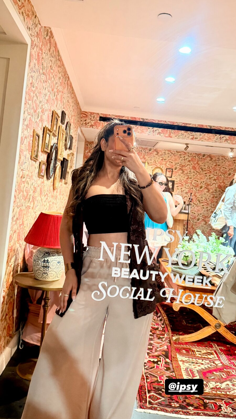 Roopsi Bansal taking a mirror selfie in a black bandeau top, cream trousers, and a burgundy velvet vest inside the IPSY Social House
