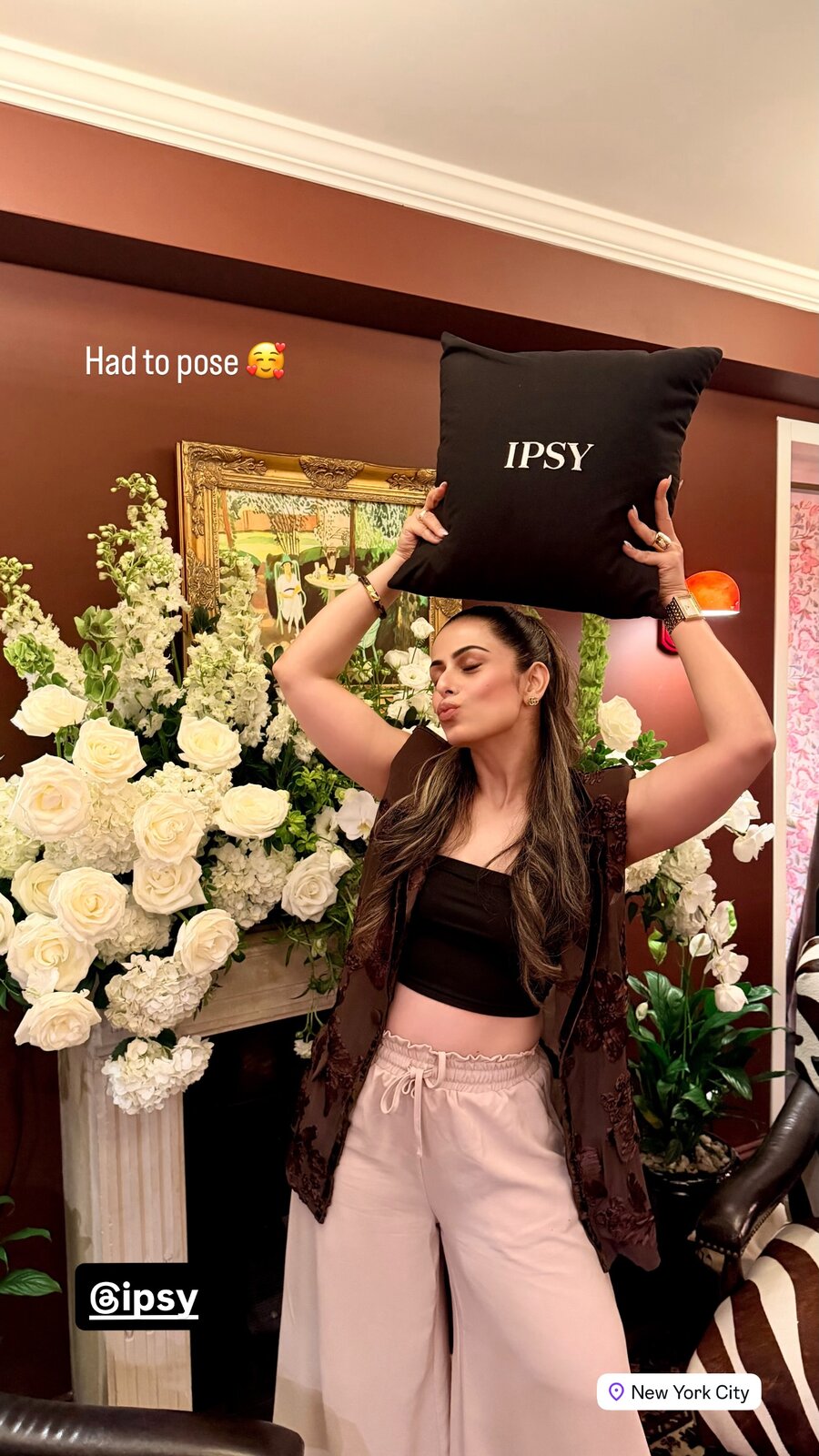 Roopsi Bansal posing with an IPSY throw pillow above her head in front of a wall of white roses and hydrangeas