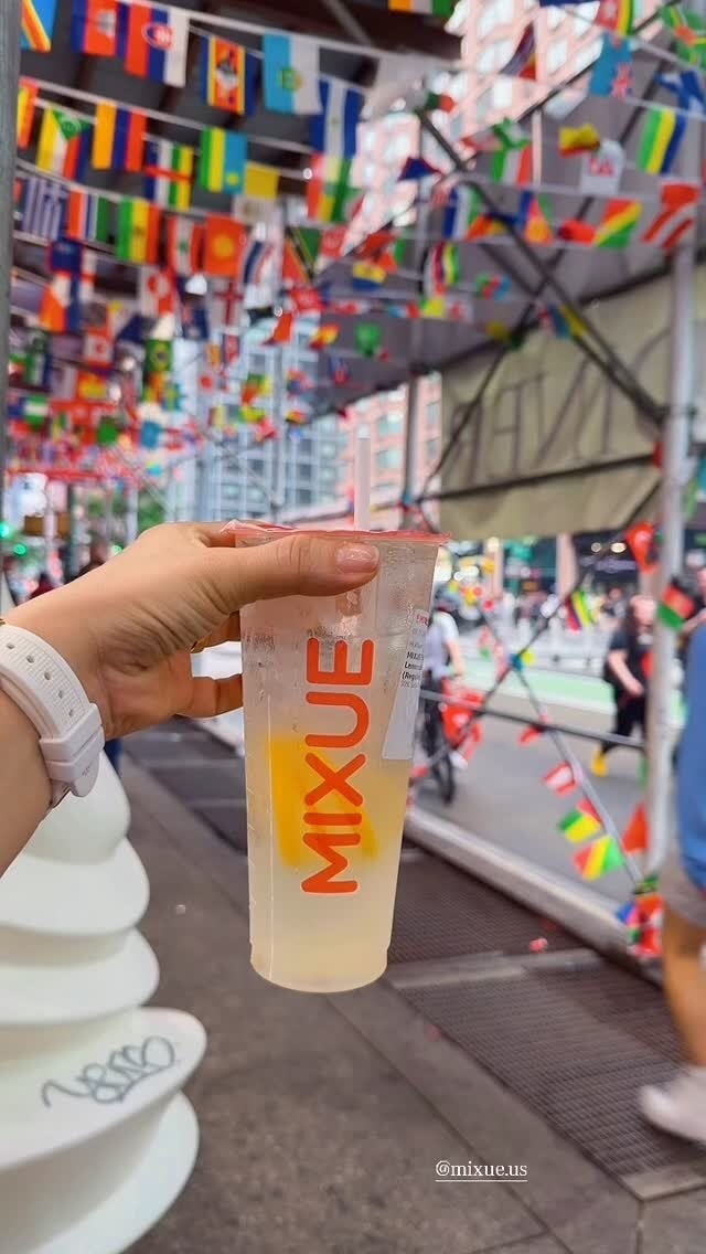 A hand holding a Mixue drink cup on a street decorated with international flags