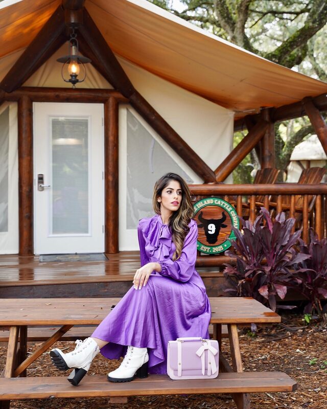 Roopsi Bansal in a purple ruffled long-sleeve dress and white lace-up boots, seated on a picnic table outside a glamping cabin numbered nine at Westgate River Ranch