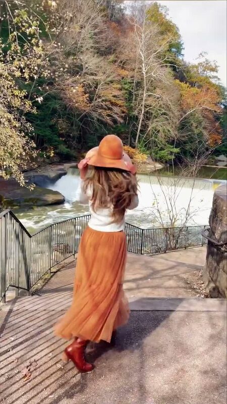 Roopsi Bansal from behind in a burnt-orange tulle maxi skirt and matching floppy hat, facing a waterfall along a fall-colored riverbank in Pittsburgh