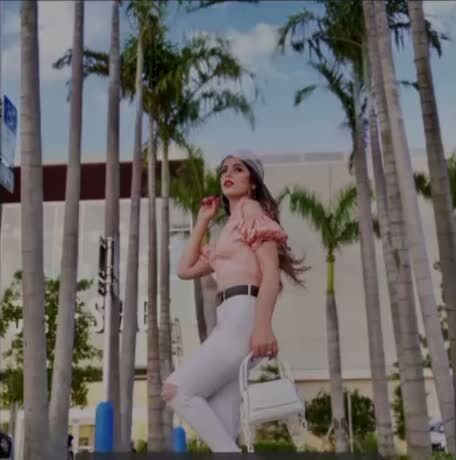 Roopsi Bansal walking through a row of palm trees in a pink ruffled off-shoulder top, white distressed jeans, white cap and white bag, filming a trending reel
