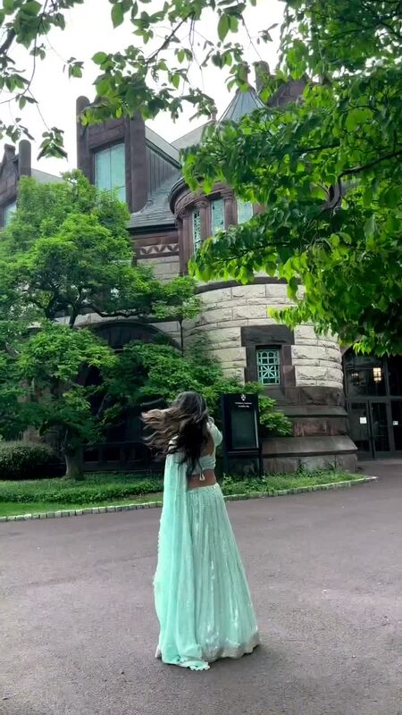 Roopsi Bansal in a mint green sequined lehenga twirling outside a grand stone building, hair blowing in the wind