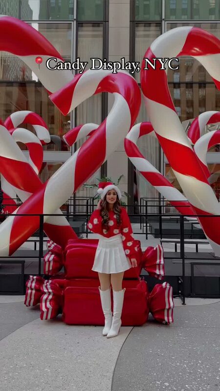 Roopsi Bansal in a red Santa-inspired sweater and white skirt beside a giant candy cane display on 6th Avenue, NYC