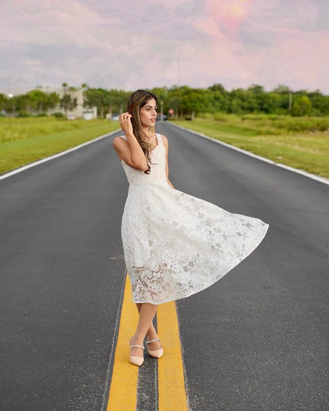 Roopsi Bansal in a white floral lace midi dress with bejeweled Mary Jane heels, standing in the middle of an empty two-lane road stretching toward the horizon