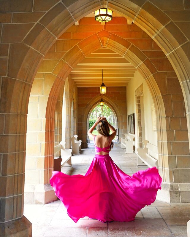 Roopsi Bansal from behind in a hot pink open-back gown, arms raised overhead, walking through a Gothic stone colonnade at The Church of Bethesda-by-the-Sea