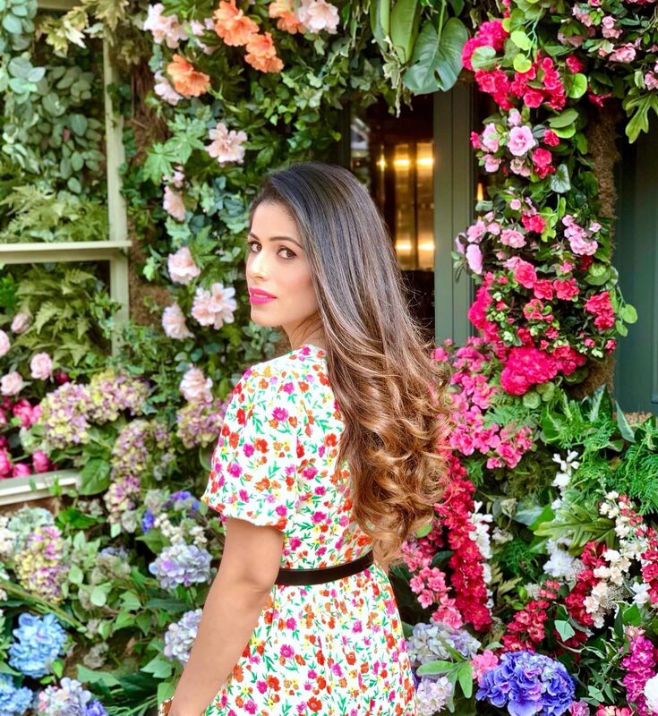 Roopsi Bansal in a floral wrap dress, looking back over her shoulder in front of a wall covered in pink and purple flowers