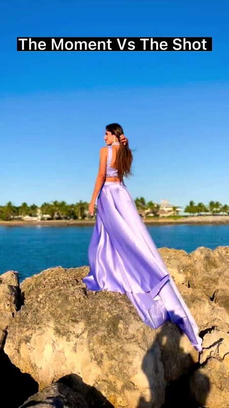 Roopsi Bansal in a lilac satin two-piece skirt set, standing on rocks along a Florida waterway with waterfront homes across the water