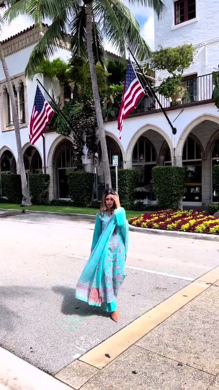 Roopsi Bansal in a turquoise Kani-woven ethnic set with a flowing dupatta, walking along Worth Avenue's palm-lined arcades with American flags overhead