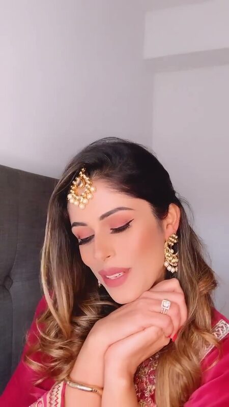 Close-up of Roopsi Bansal in a pink and red embroidered outfit with gold maang tikka and earrings, resting her chin on her hand