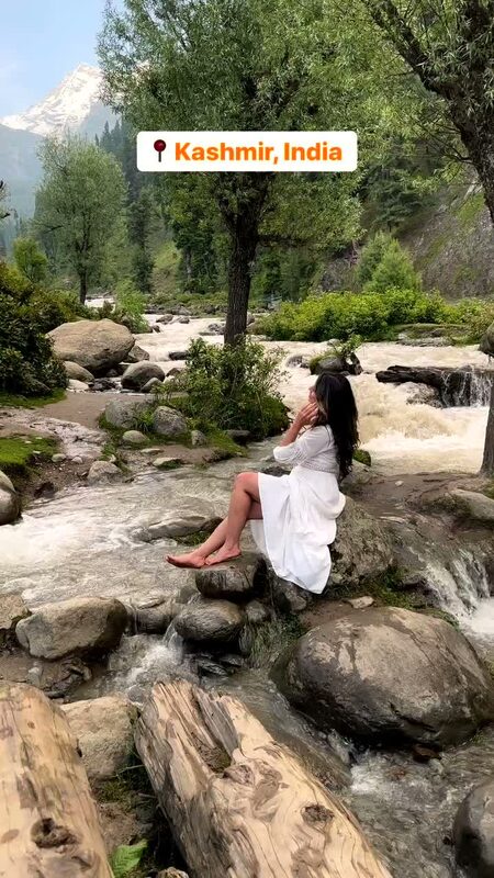 Roopsi Bansal in a white dress sitting on rocks beside a rushing glacial stream in Kashmir, with snow-capped mountains in the distance