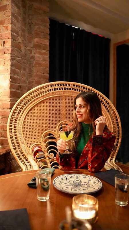 Roopsi Bansal seated in a woven peacock chair holding a cocktail at Kebab Aur Sharab restaurant in Manhattan