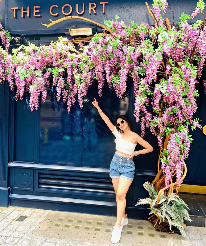 Standing beneath a wisteria-covered pub storefront called The Court, pointing up at the flowers, wearing a white one-shoulder crop top and denim shorts