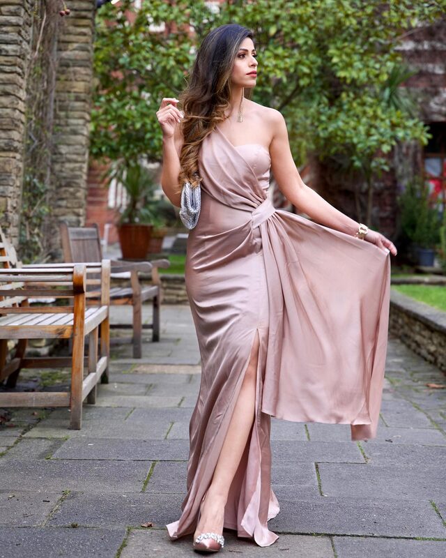 Roopsi Bansal in a blush one-shoulder satin gown with a draped thigh slit, holding a silver beaded clutch in a garden courtyard