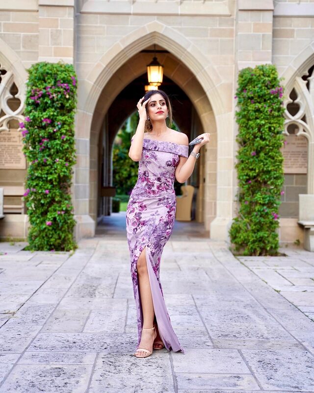 Roopsi Bansal in an off-shoulder lilac floral-print gown with a thigh-high slit, standing under a Gothic stone archway flanked by bougainvillea