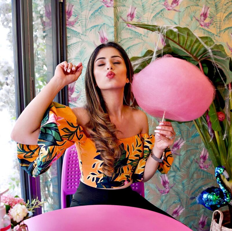 Roopsi Bansal in a navy ruffle-sleeve top, blowing a kiss beside a large pink cotton candy cloud at a table set with desserts, in front of a botanical-print wall