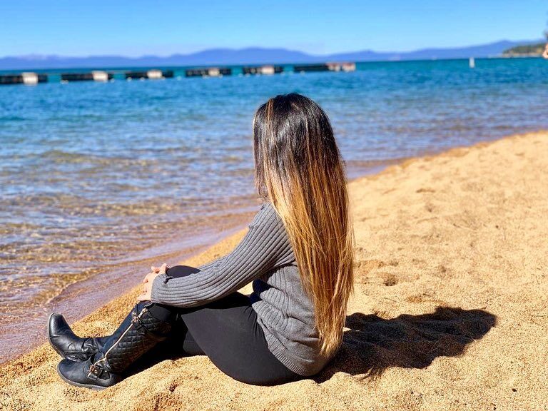 Sitting on the sandy shore of Lake Tahoe in a grey sweater and black boots, looking out at the turquoise water and mountains
