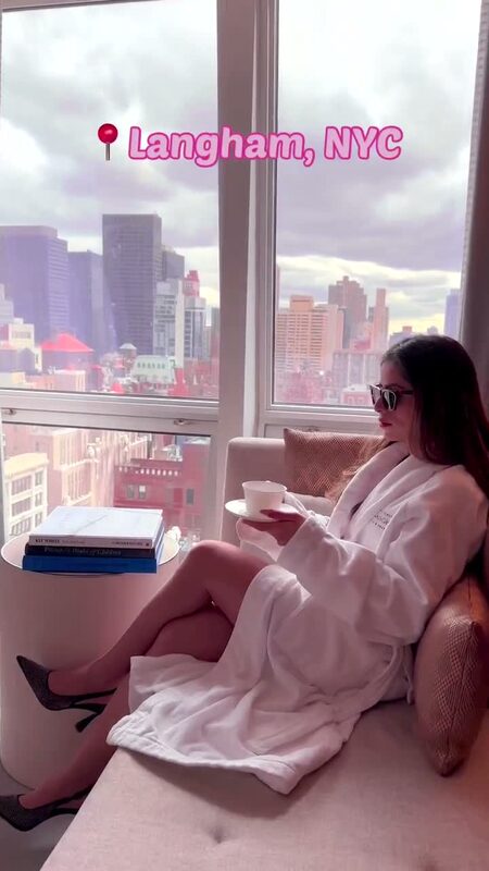 Roopsi Bansal in a white hotel robe sipping coffee by a window overlooking the New York City skyline at The Langham