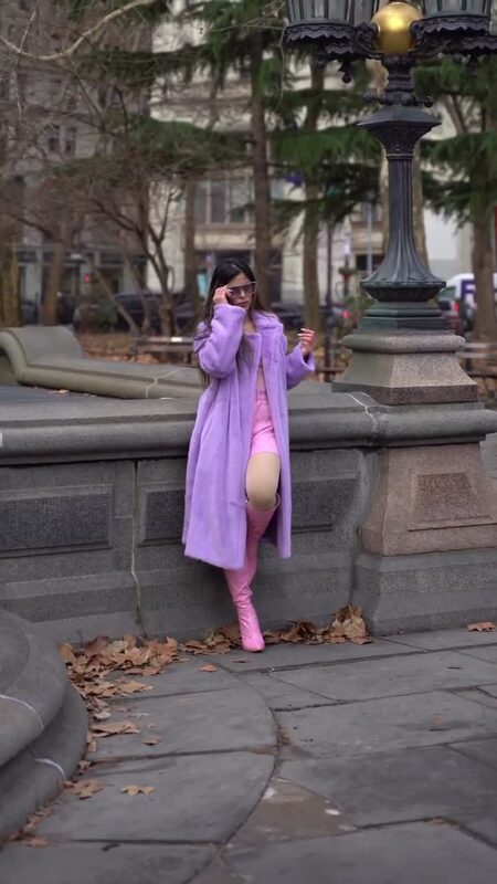 Roopsi Bansal in a lavender faux fur coat and pink boots leaning against a stone fountain wall near City Hall Park
