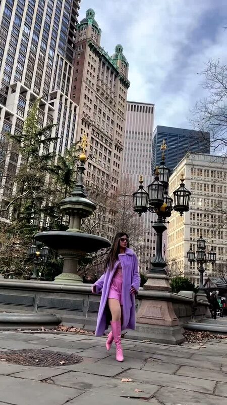 Roopsi Bansal in a lavender faux fur coat and pink boots standing by an ornate fountain near City Hall Park in New York