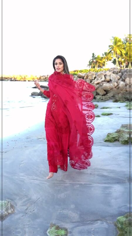 Roopsi Bansal in a red embroidered salwar suit with a sheer red scalloped dupatta caught mid-twirl, walking barefoot on a Florida beach at sunset