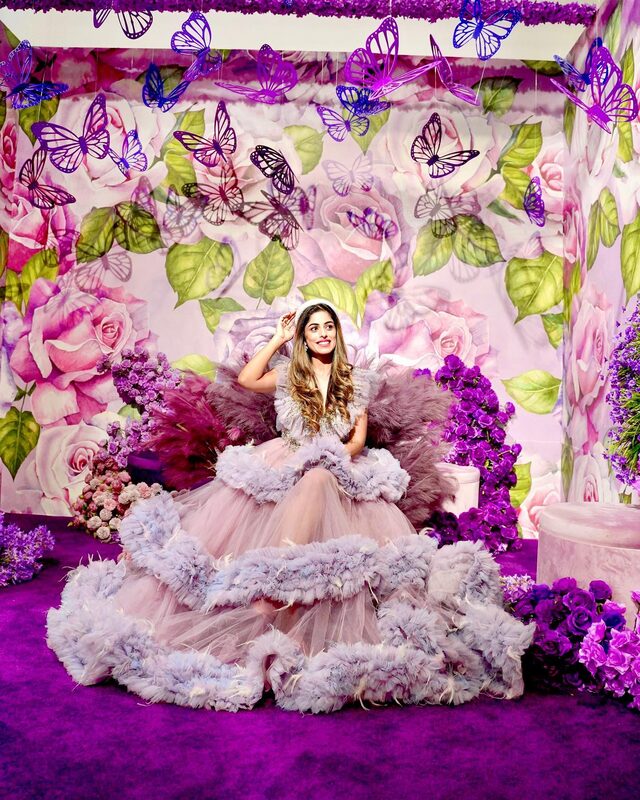 Roopsi Bansal in a lavender feathered tiered ball gown, seated in a room covered in painted roses and hanging purple butterflies at a floral installation
