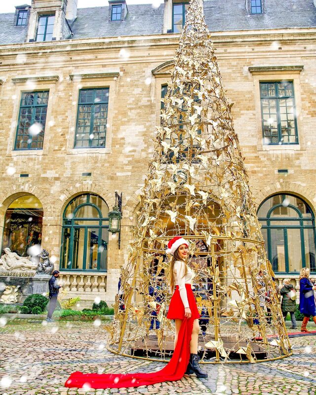 Roopsi Bansal in a Santa hat, white sweater and long red skirt with a dramatic train, standing beneath a golden origami-crane Christmas tree in a historic courtyard in Brussels