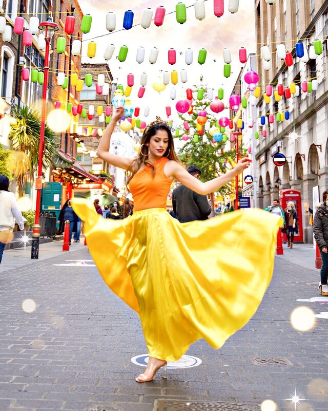 Roopsi Bansal in an orange halter top and flowing yellow satin skirt, mid-twirl on a Chinatown London street strung with colorful paper lanterns