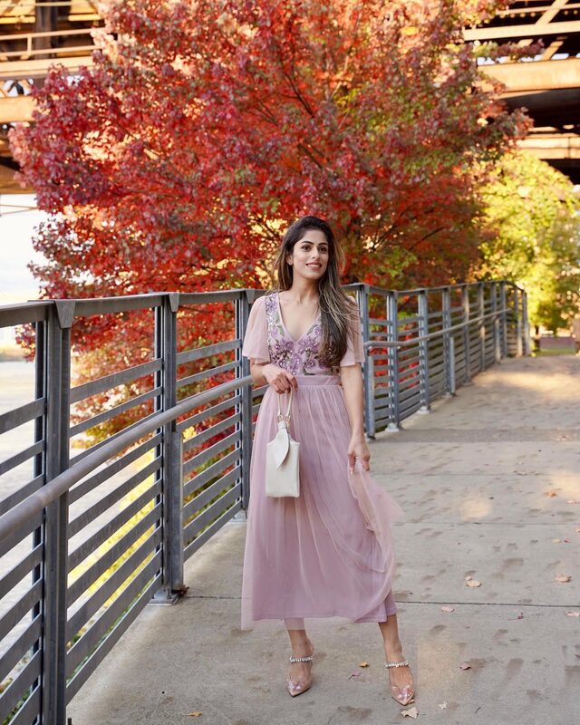 Roopsi Bansal in a dusty pink floral embroidered midi dress with flutter sleeves, holding a white handbag, on a footbridge lined with a tree in full red fall color