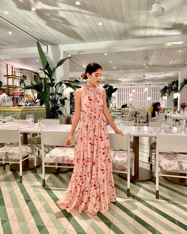 Roopsi Bansal in a pink floral print halter maxi dress and statement flower earrings, standing in the dining room at Honeybelle restaurant at PGA National Resort
