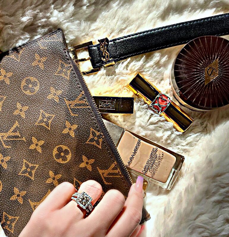 A flat-lay on a faux fur rug of a Louis Vuitton monogram wallet, a YSL belt and lipstick, a Charlotte Tilbury compact, and a diamond ring