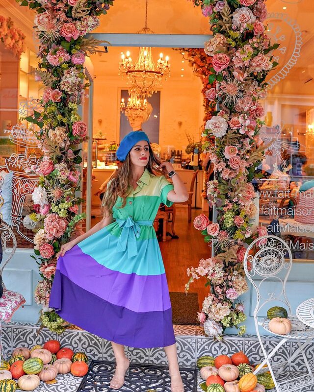 Roopsi Bansal in a green, teal and purple colour-block dress and blue beret, standing in a floral archway surrounded by pumpkins outside a cafe