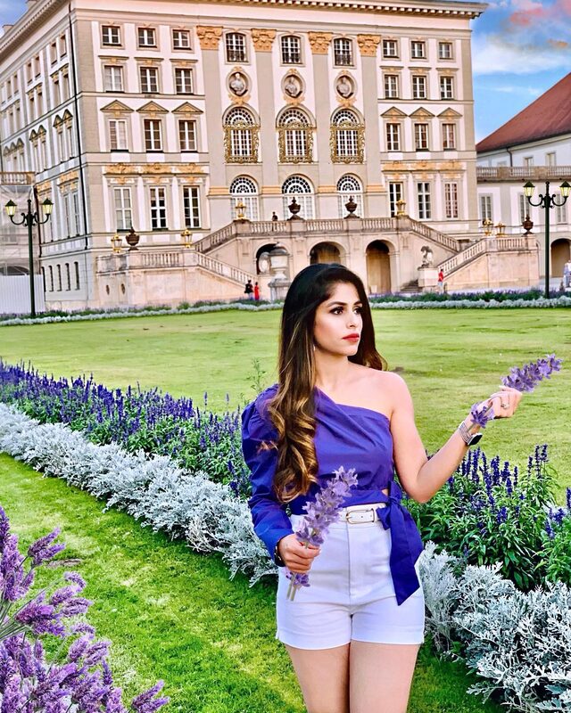 Roopsi Bansal in a purple one-shoulder top and white shorts, holding lavender stems in front of an ornate palace facade with a bed of purple salvia flowers