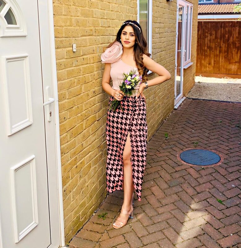 Walking down a brick side passage between houses holding a bouquet of flowers, wearing a pink off-shoulder ruffle top and a red and white houndstooth wrap skirt