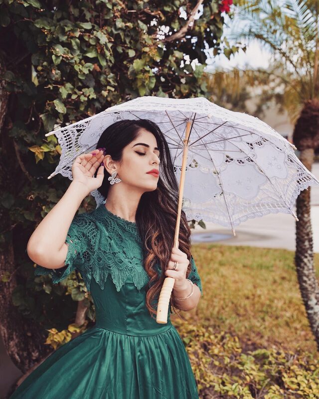 Roopsi Bansal in an emerald green lace-trim dress and butterfly earrings, holding a white lace parasol in front of a garden hedge