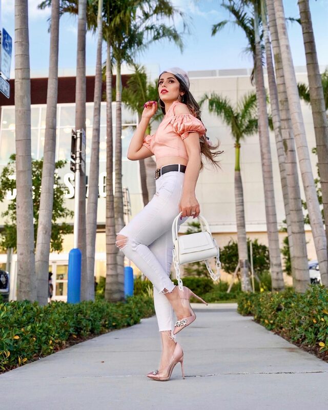 Roopsi Bansal in a peach off-shoulder puff-sleeve top and ripped white jeans, carrying a white chain-strap handbag on a palm-lined sidewalk in Miami