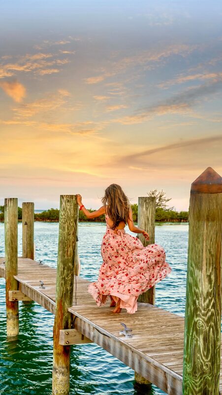 Roopsi Bansal from behind in a pink floral maxi dress, walking along a wooden dock over turquoise water at sunset at Hawks Cay Resort