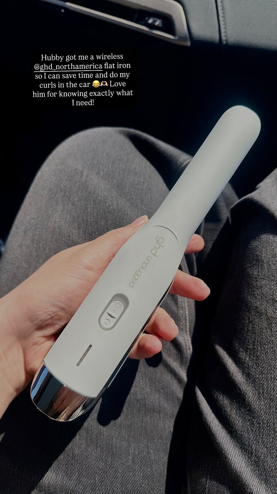 A hand holding a wireless GHD flat iron on a lap inside a car
