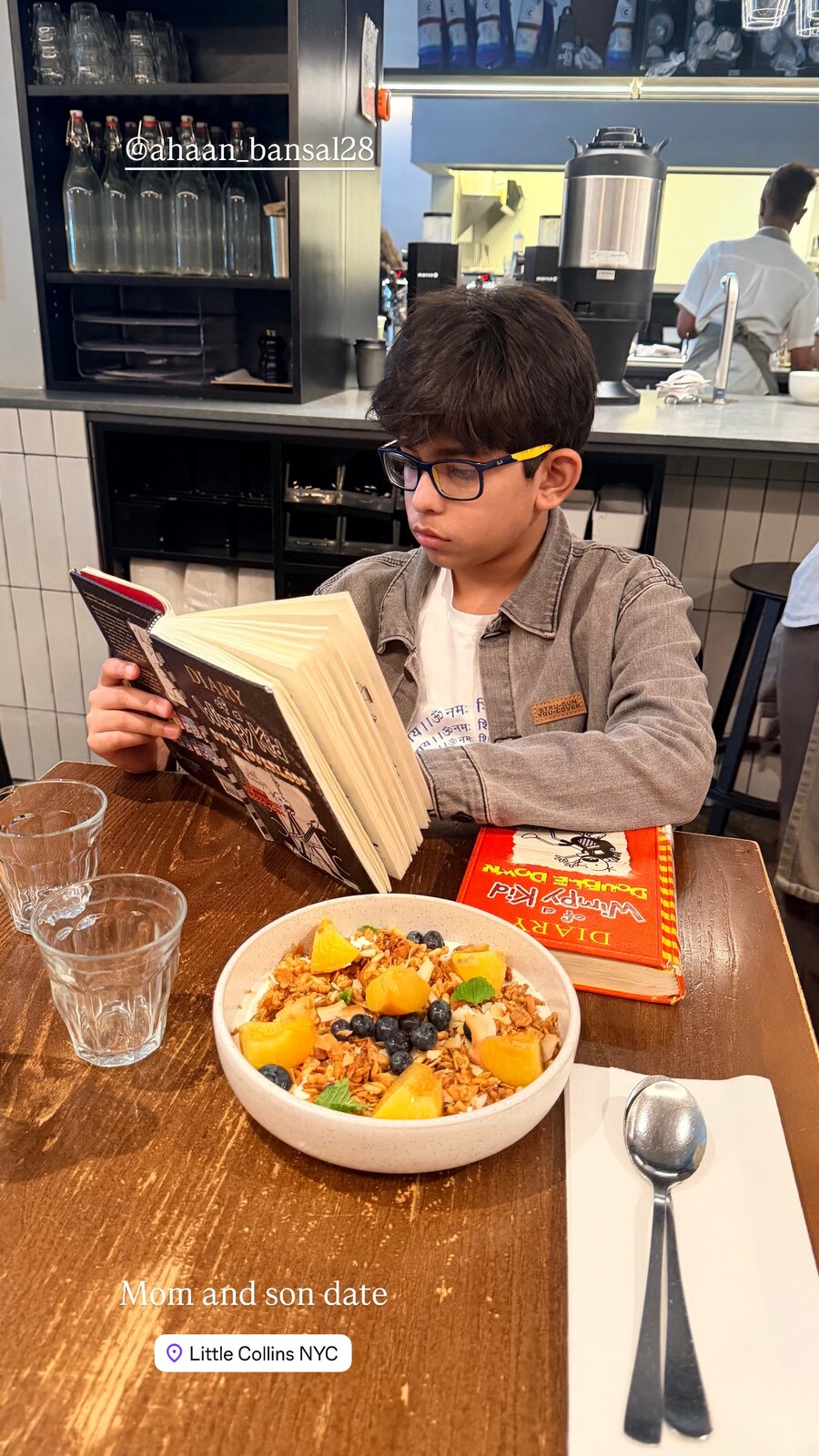 A boy reading Diary of a Wimpy Kid at a table at Little Collins NYC, with a bowl of granola, yogurt, peaches and blueberries in front of him