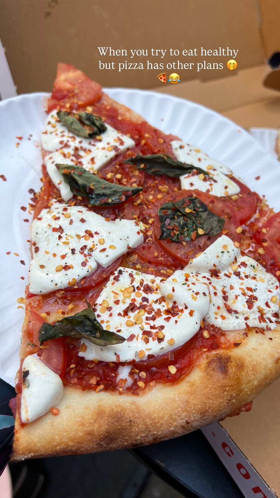 A slice of margherita pizza topped with mozzarella, tomato, basil and chili flakes on a paper plate