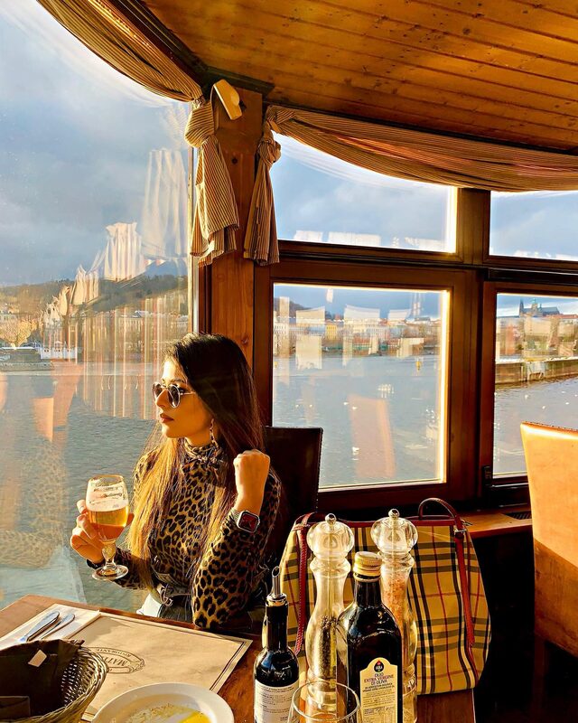 Sitting at a table on a restaurant boat on the river, wearing a leopard-print top, holding a glass of beer, with a Burberry check bag beside the window and the river view behind