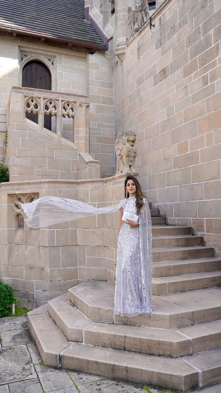 Roopsi Bansal in a silver sequined caped gown from Frock and Frill, holding a white clutch from Avandlina, standing on the stone steps of a historic building beside a carved lion statue