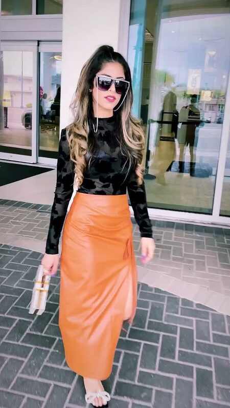 Roopsi Bansal in a black mesh top and orange faux-leather midi skirt, walking past mirrored storefronts near Hard Rock Hotel Daytona Beach